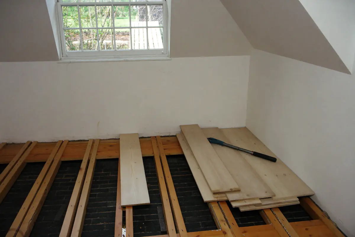 Zolderkamer van een oud huis waar de grenen vloerplanken deels zijn weggehaald, met de houten balken zichtbaar en de losse planken gestapeld