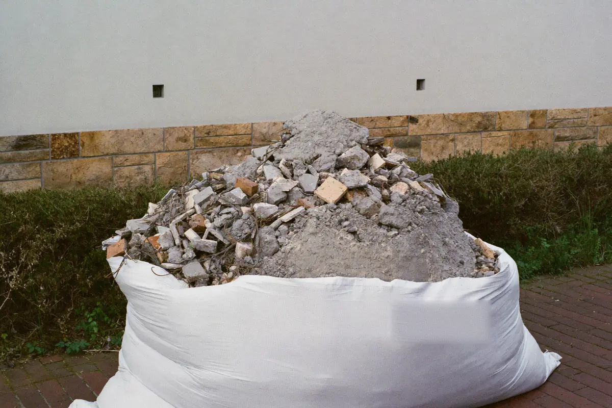 Gevulde witte big bag met gebroken tegels en dekvloerpuin op een klinkeroprit voor een huis met een plint van gele natuursteen